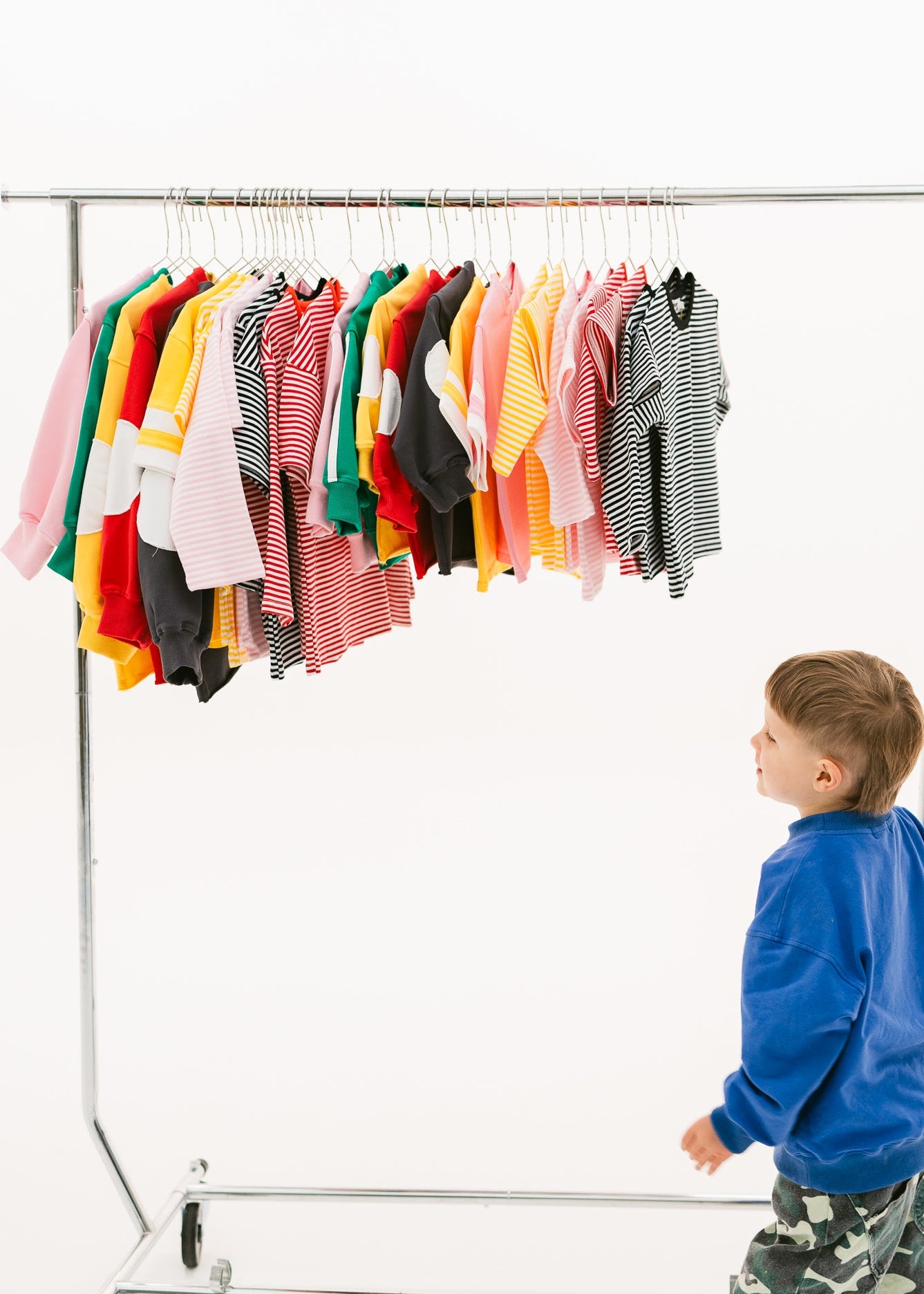 A young child in a blue sweatshirt and camouflage pants walks beside a metal rolling clothing rack filled with colorful children's shirts, photographed on a clean white studio background.
