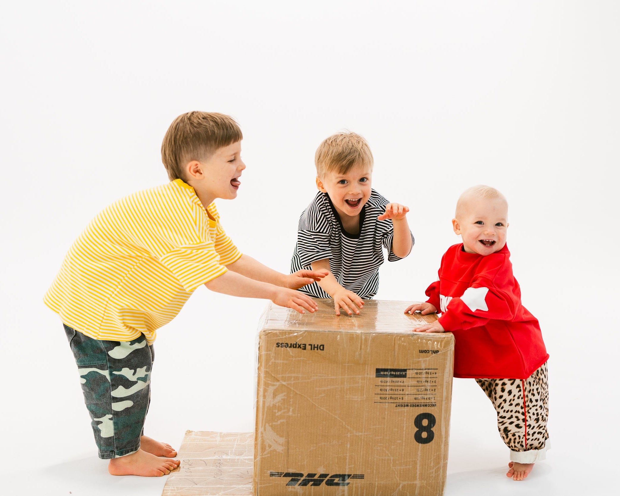Three children play with a large cardboard box on a white studio background. One child in a yellow striped shirt pushes the box, while another in a gray striped shirt and a baby in a red outfit watch and smile.