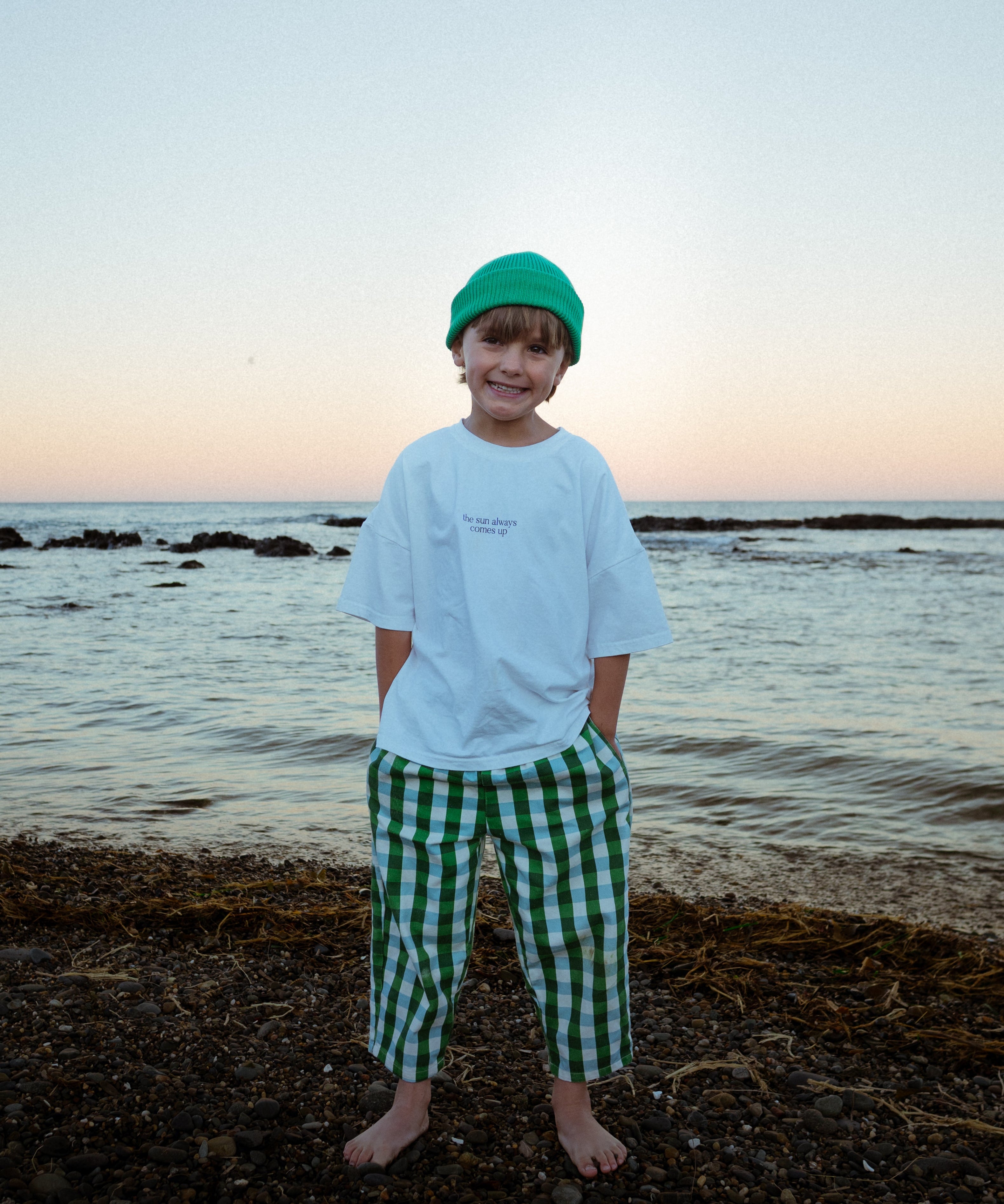 Child standing on a beach at sunset wearing a white shirt and green checkered pants.