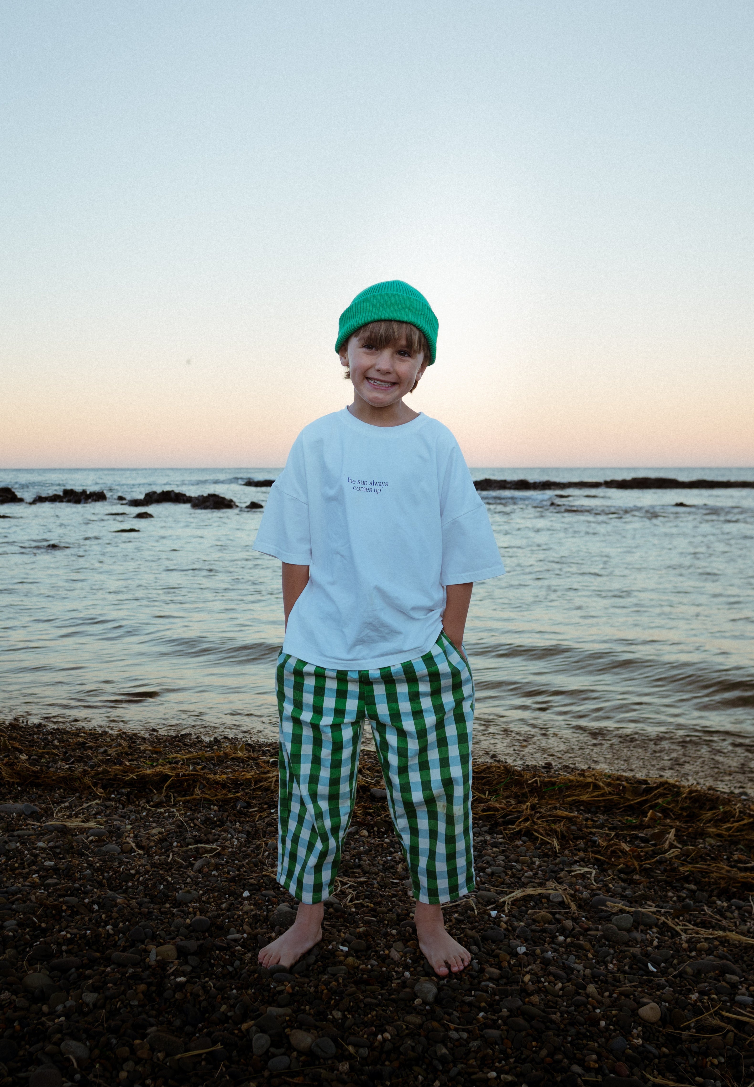 Child standing on a beach at sunset wearing a white shirt and green checkered pants.