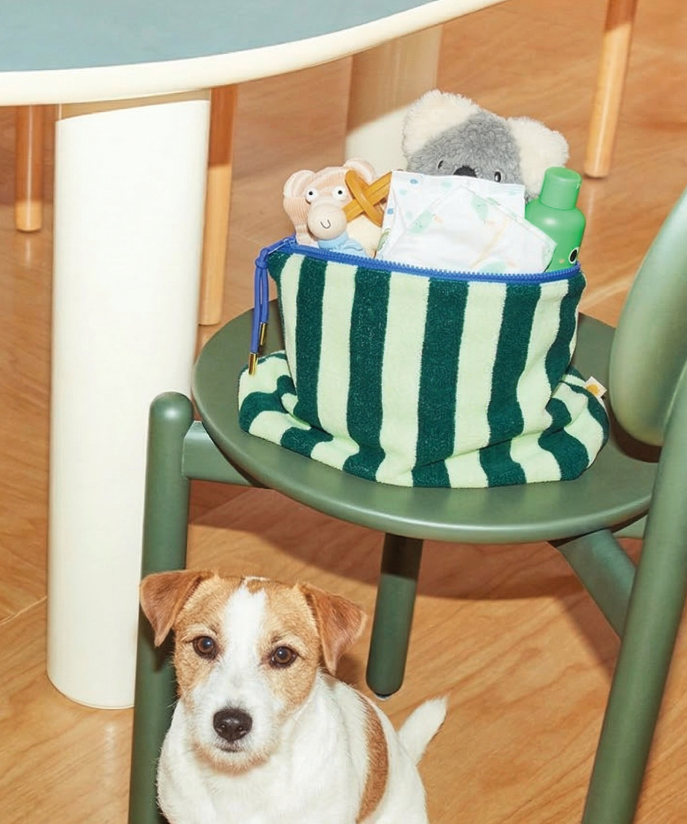 Sonny Label mint green stripe baby pouch on a sage green chair, styled with baby toys and a little white dog nearby.