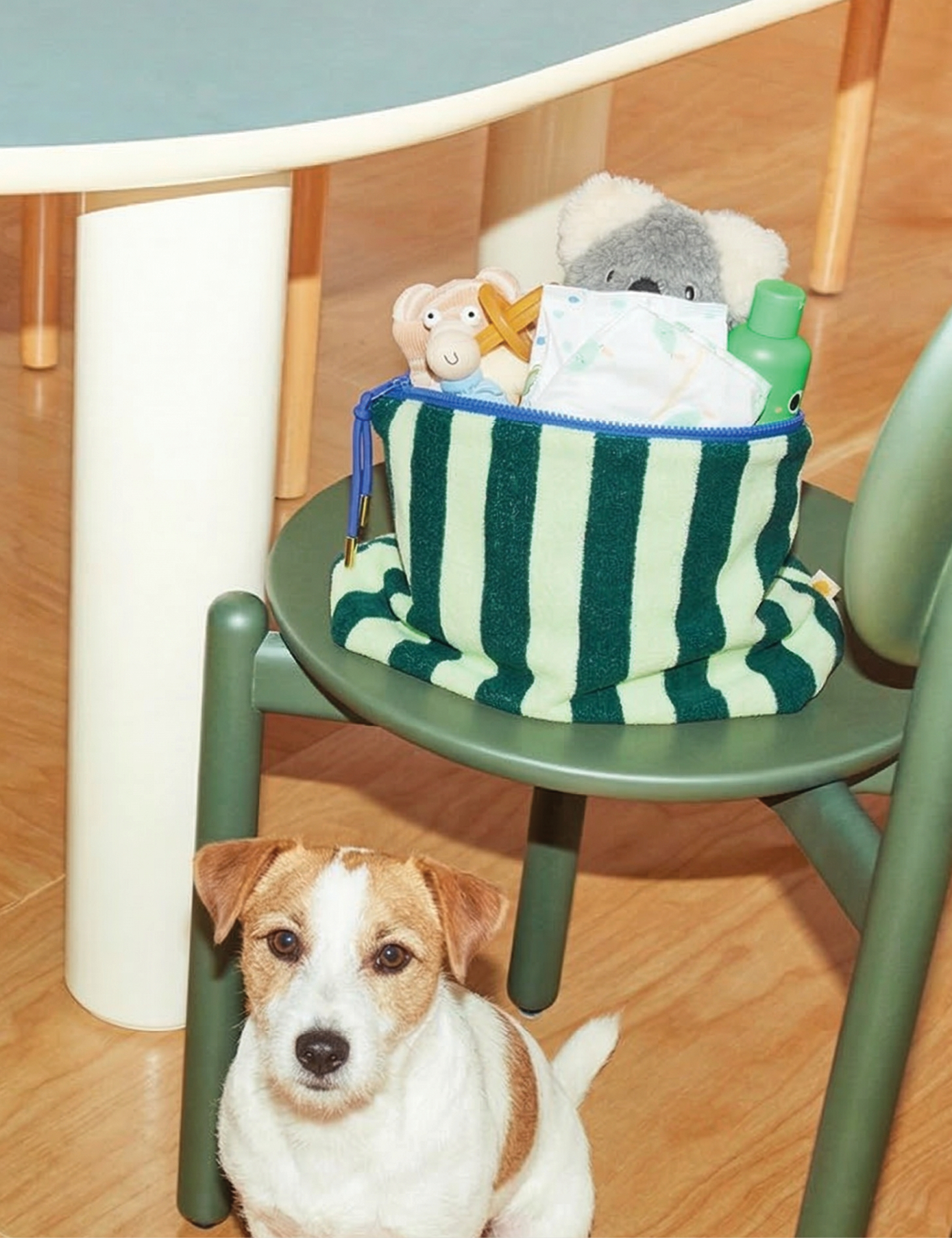 Sonny Label mint green stripe baby pouch on a sage green chair, styled with baby toys and a little white dog nearby.