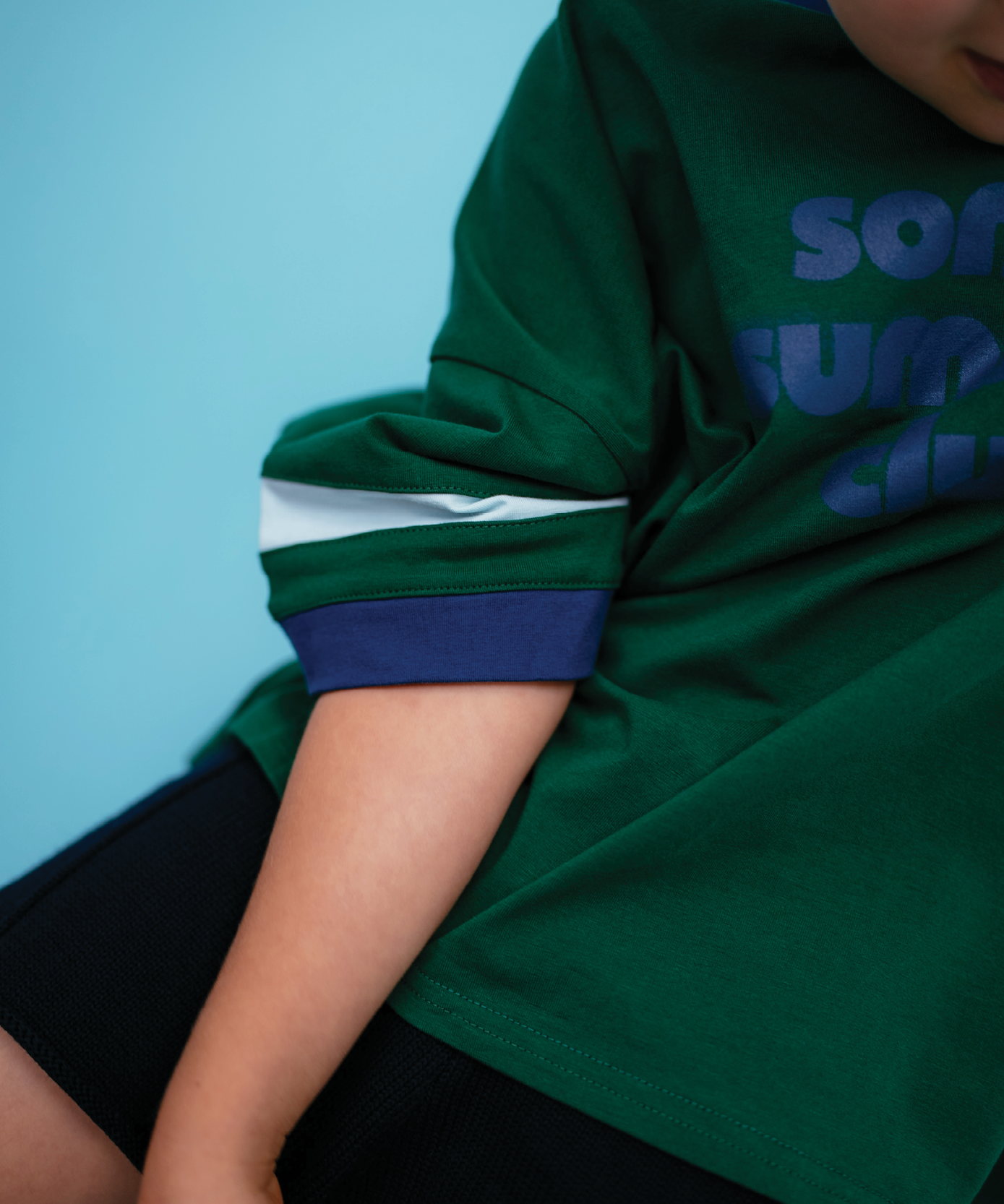 Shoulder detail of a Sonny Label Summer25 forest green "Sonny Summer Club" sport tee on a sky blue background.