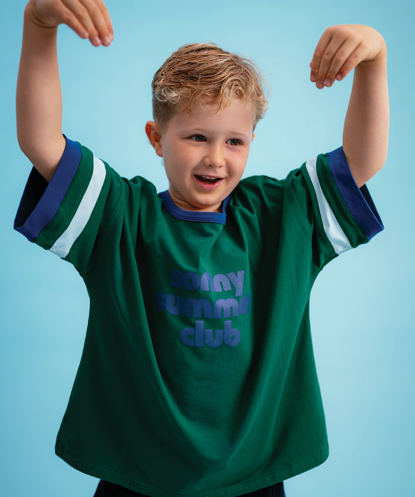 Boy wearing Sonny Label Summer25 "Sonny Summer Club" forest green sport tee with arms raised on a sky blue background.