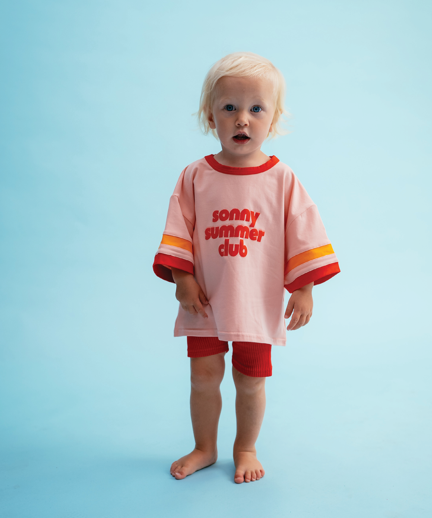 Child wearing Sonny Label Summer25 beach poncho and bikies on a sky blue background.