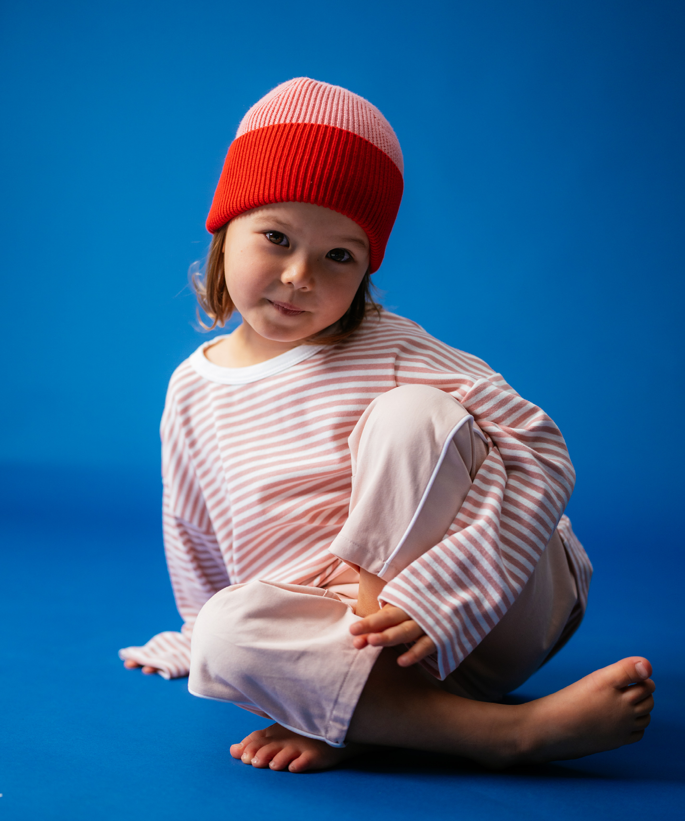Girl in Sonny Label Circus26 pink and white stripe long-sleeve tee and floss slouch pants with red beanie, sitting cross-legged on a vivid blue backdrop.