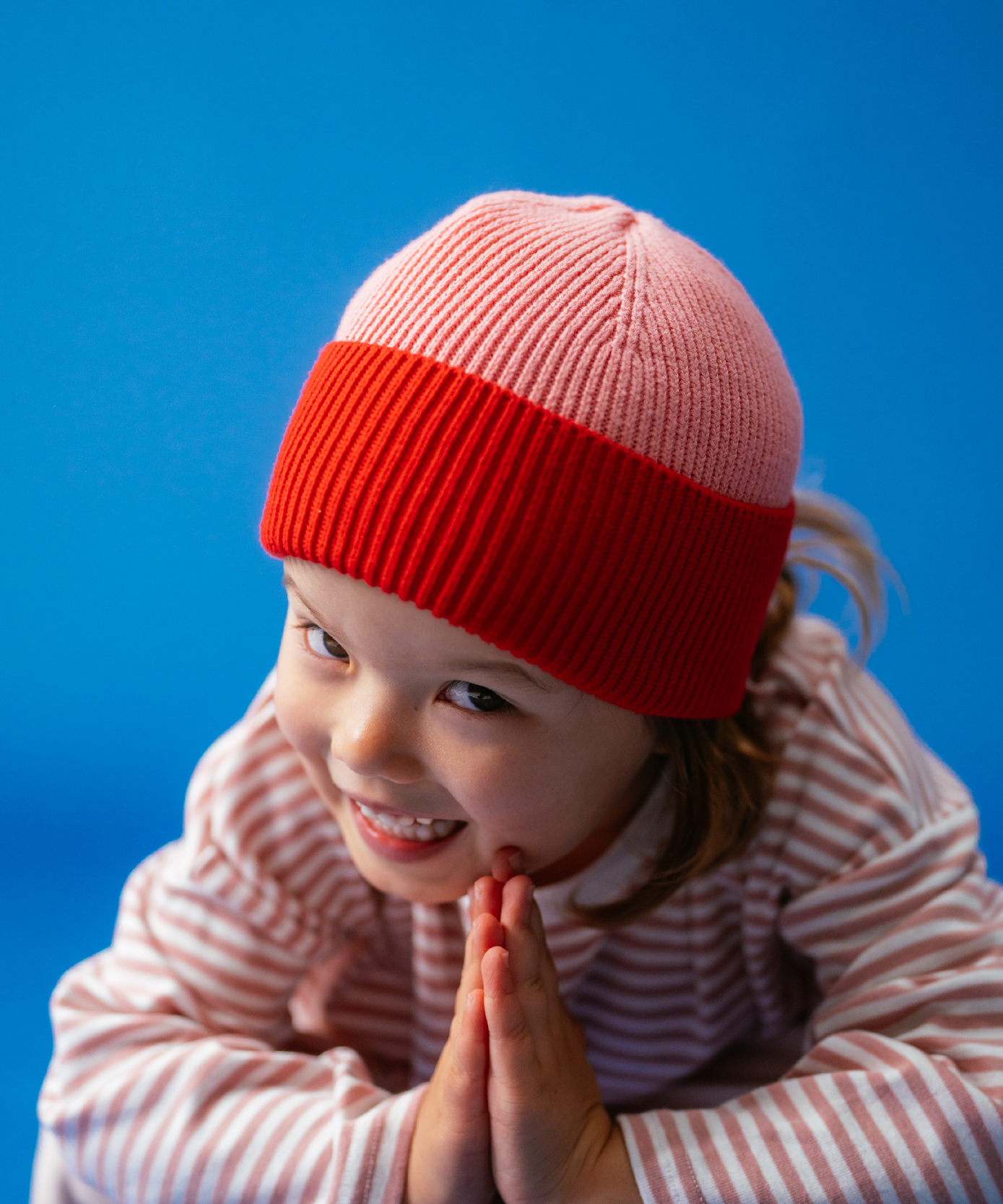 Close-up of a girl in Sonny Label Circus26 pink stripe LS and floss pants, hands in a prayer pose on a blue backdrop.