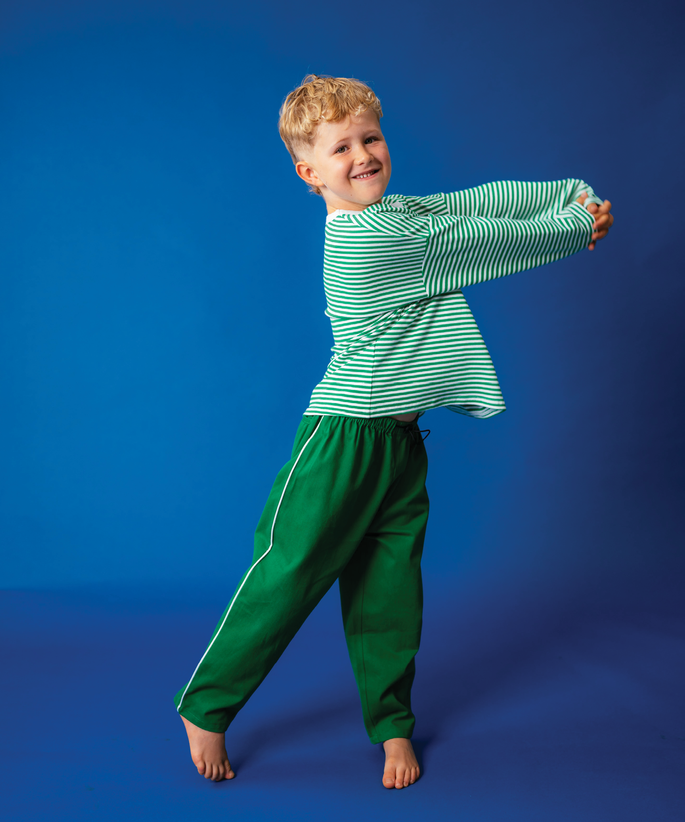 Child in Sonny Label Circus26 stripe tee and forest green sport pants on a deep blue background.