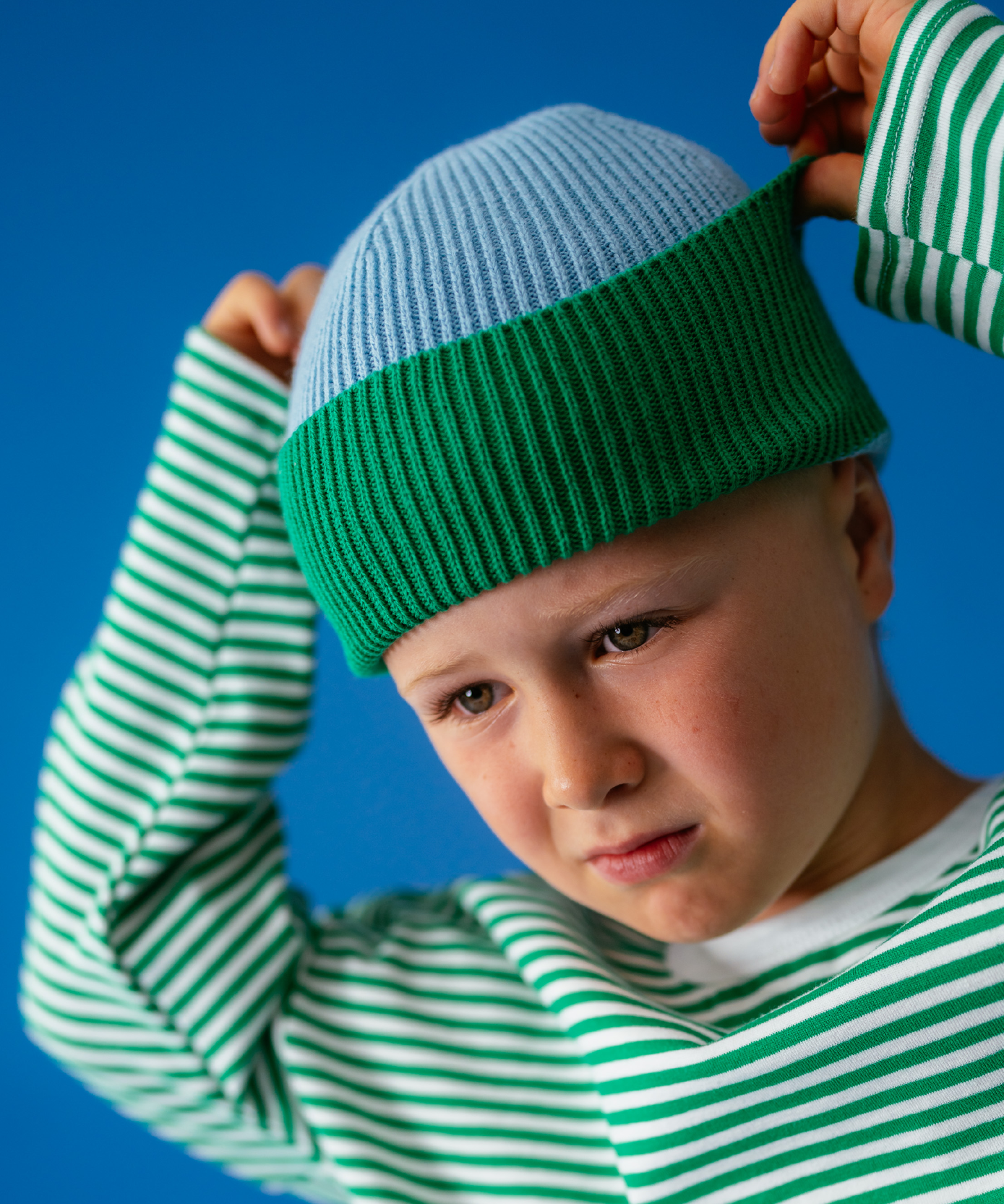 Boy in Sonny Label Circus26 green and white stripe long-sleeve tee with sky and green beanie, arms raised on a blue backdrop.