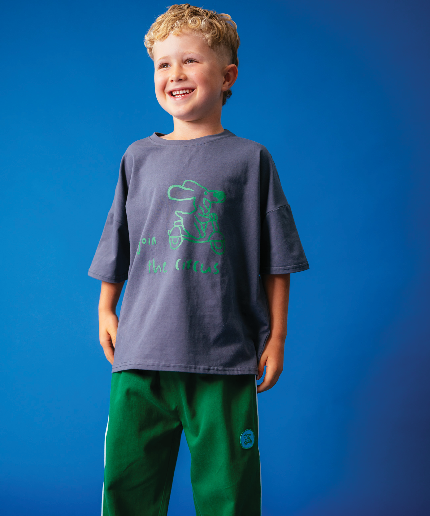 Child wearing Sonny Label Circus26 sport tee and floss pink pants on a vivid blue background.
