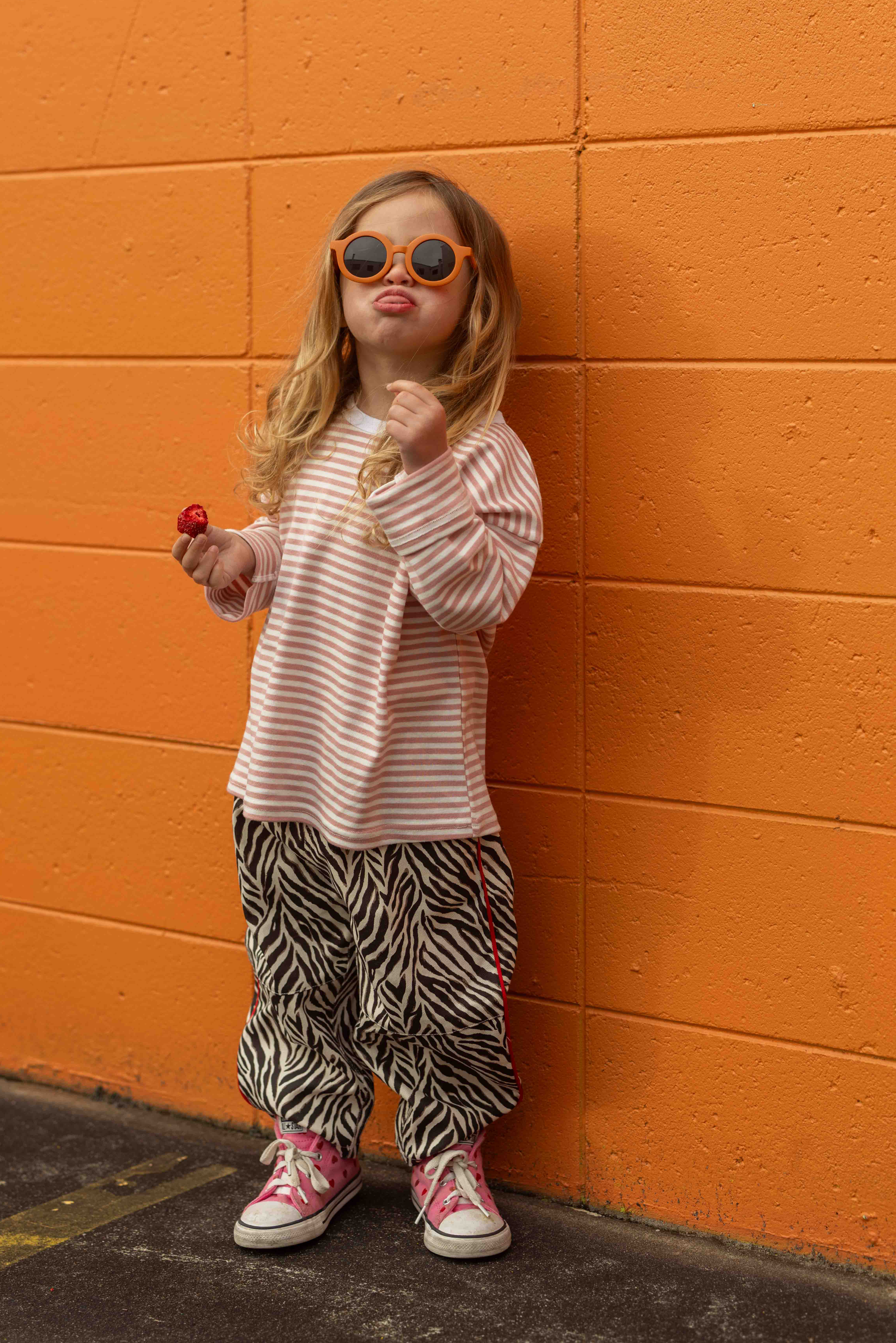Girl in Sonny Label Safari AW25 zebra print pants and pink stripe long-sleeve tee with orange sunglasses, leaning on an orange wall holding a lollipop.